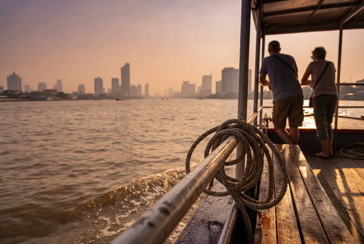 Hai hành khách tựa lan can một chiếc thuyền trên sông, nhìn qua sông Chao Phraya về phía đường chân trời thành phố mờ sương lúc hoàng hôn