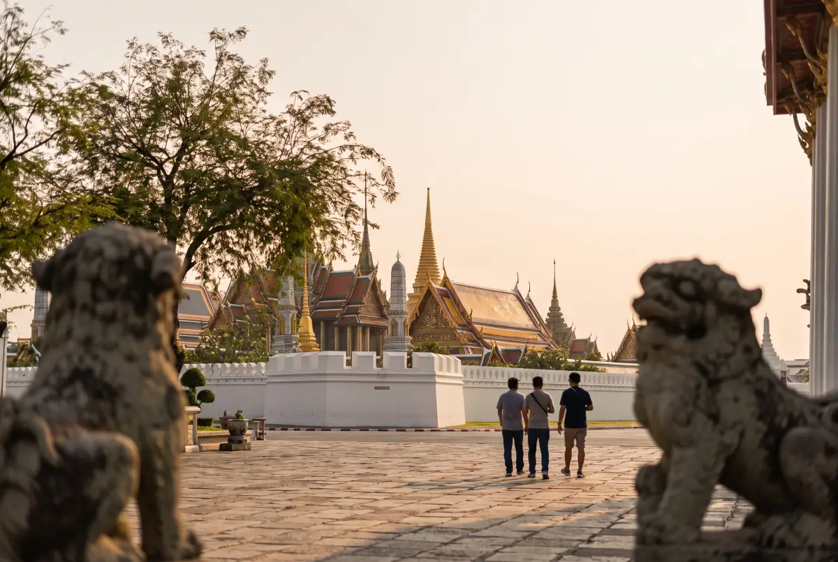 Gilded spires and tiered temple roofs rising behind a long white wall, framed by stone guardian lions in a paved courtyard