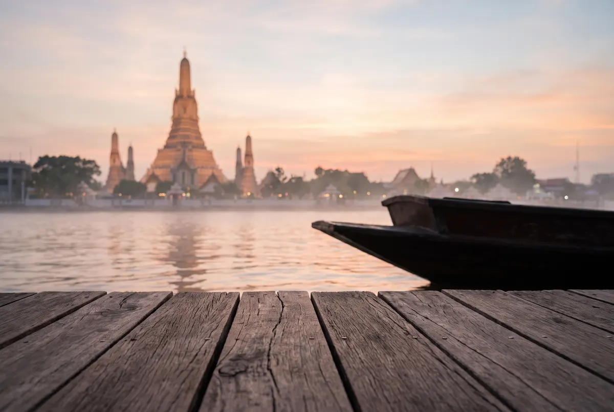 The tall riverside prang and its flanking towers seen out of focus across the water from a wooden boat deck at dawn