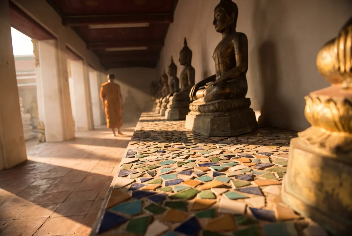 A temple cloister lined with seated bronze Buddhas above a plinth of coloured tile mosaic, a monk walking away down the sunlit corridor