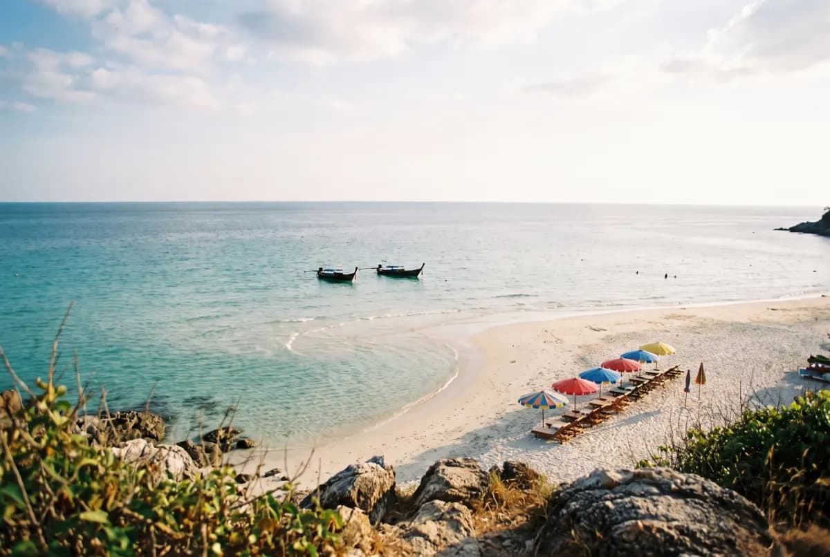 A curved white-sand beach with a row of coloured umbrellas and two longtail boats on turquoise water, seen from a rocky headland above