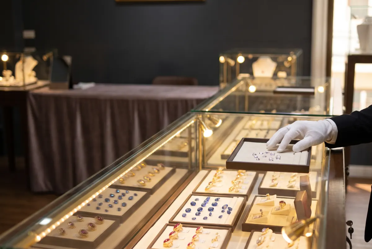 A lit glass counter of loose sapphires and gemstone rings, with a white-gloved hand setting down a tray of cut stones