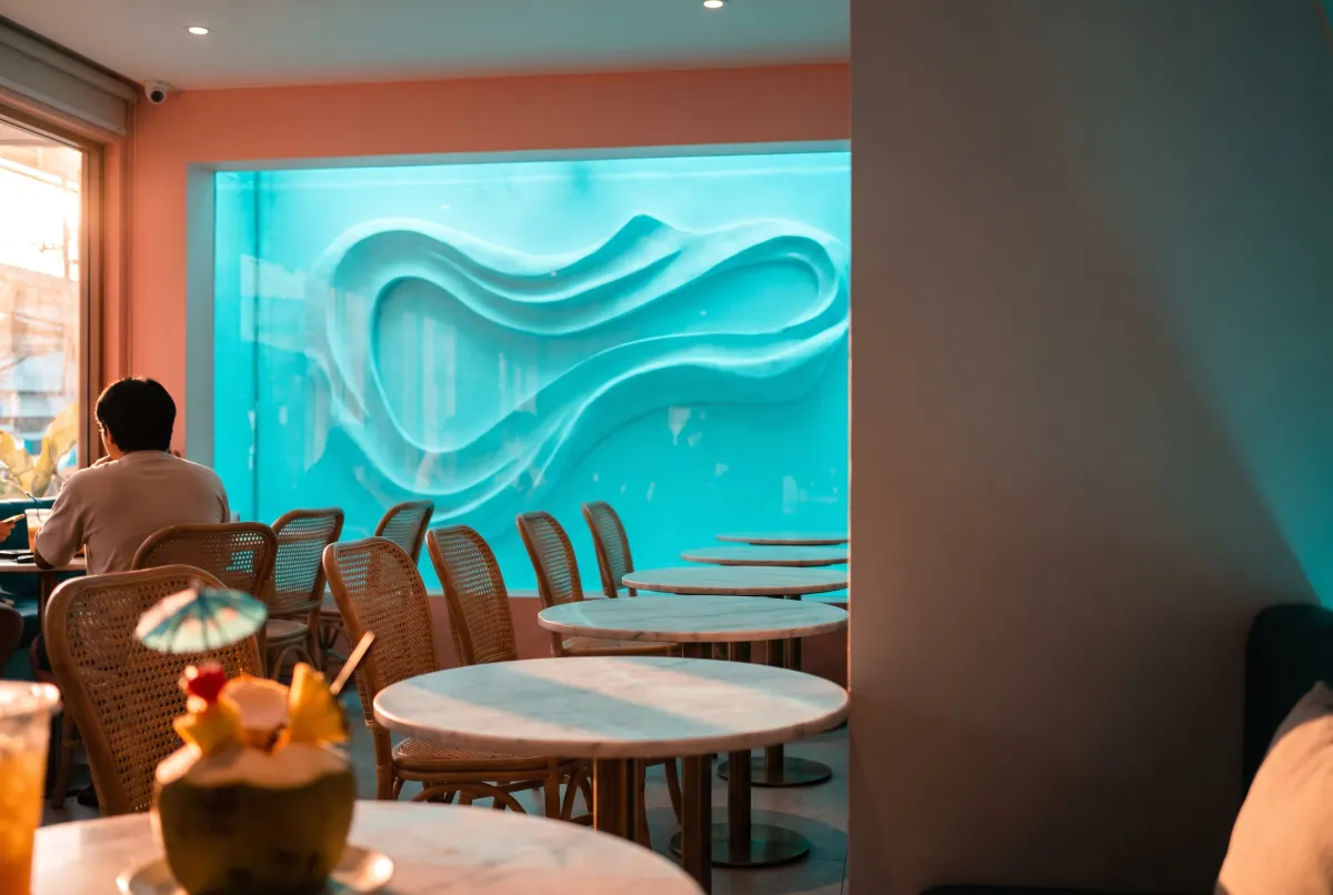 A cafe interior with rattan chairs, marble tables and a backlit turquoise wave panel, a coconut drink on the table in the foreground