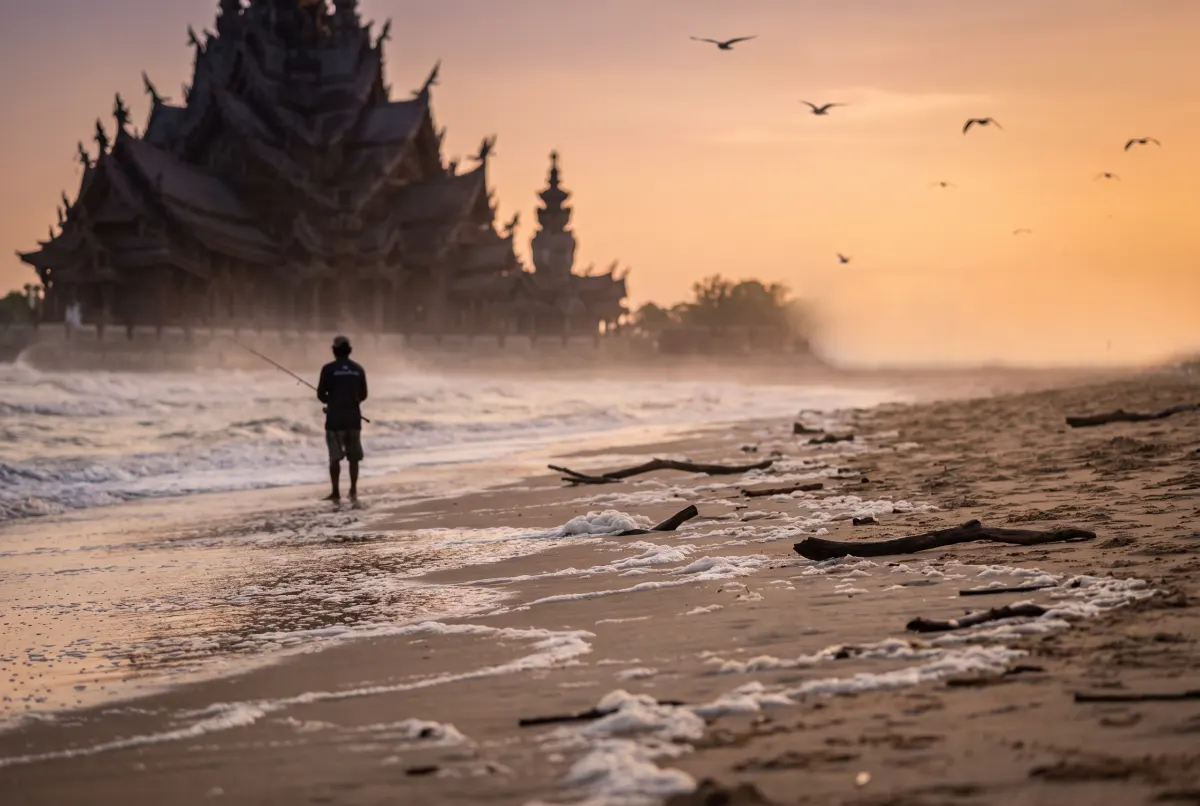 A fisherman casting from a driftwood-strewn beach at sunset, with a tiered carved wooden temple standing out of focus behind him