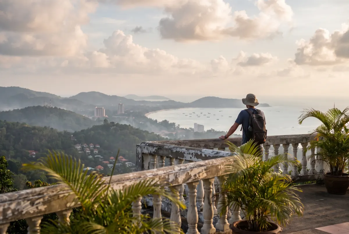 A visitor standing at a white balustrade looking out over a forested headland and a wide curving bay dotted with moored boats